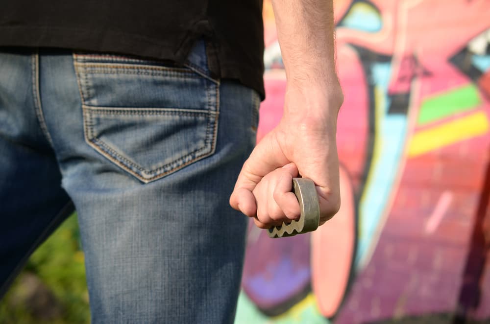 Back view of a young Caucasian man standing against a graffiti-covered brick wall in a gritty urban setting, wearing brass knuckles on his hand. The scene conveys a sense of aggression and criminal intensity.